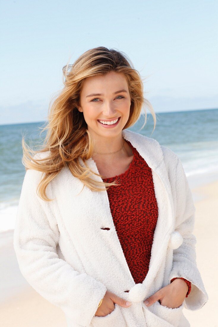 A young blonde woman on a beach wearing a red jumper and white teddy fleece jacket