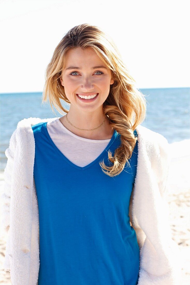 A young blonde woman on a beach wearing a blue top and a white teddy fleece jacket