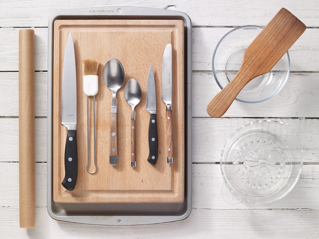 Assorted knives and spoons and a brush on a wooden board, a citrus press, a glass bowl, a spatula and baking paper
