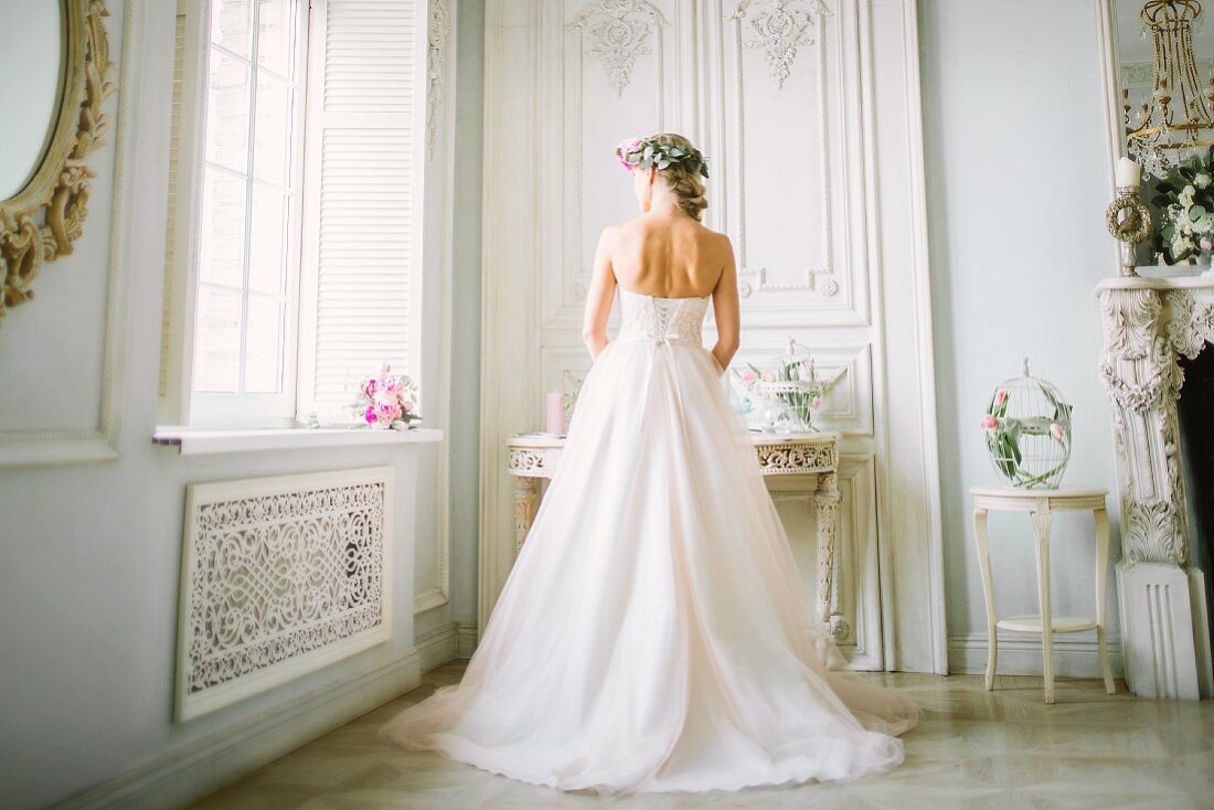 A young blonde woman wearing a wedding dress and looking out a window