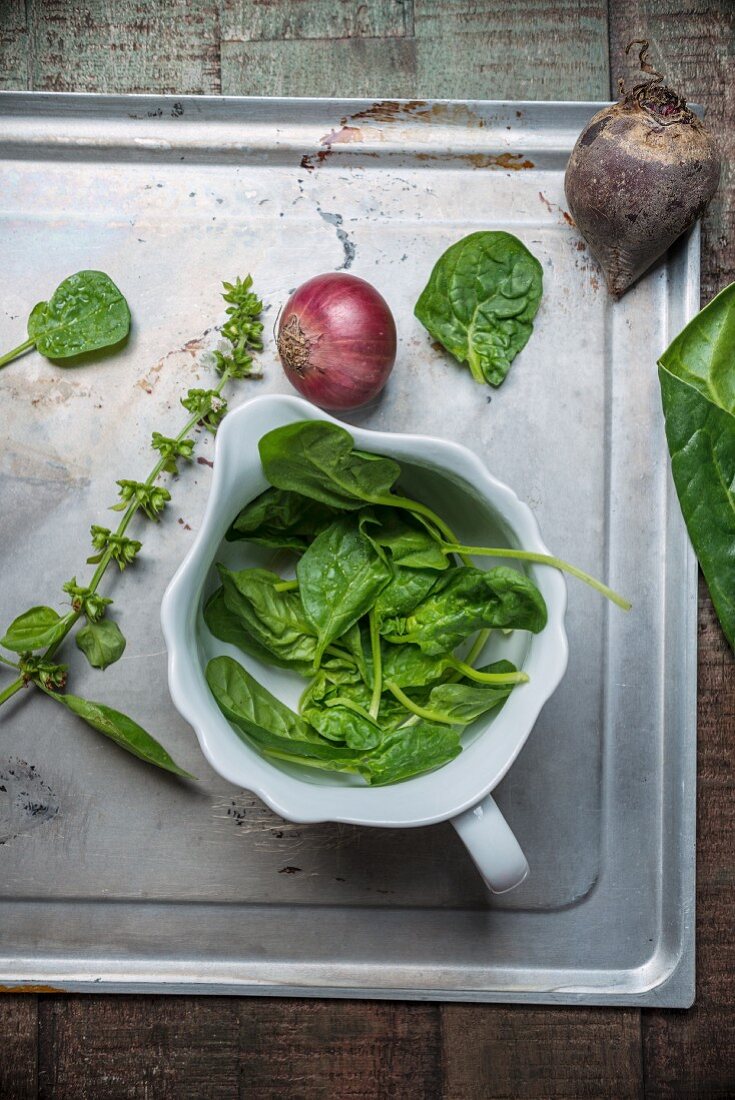Fresh spinach in a porcelain bowl on a baking tray and on a wooden surface, beetroot, basil, red onions and chard