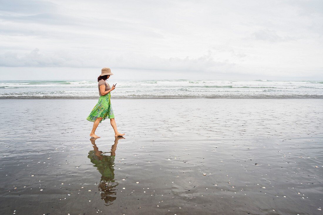 A woman in a green summer dress walking along the beach holding her mobile phone