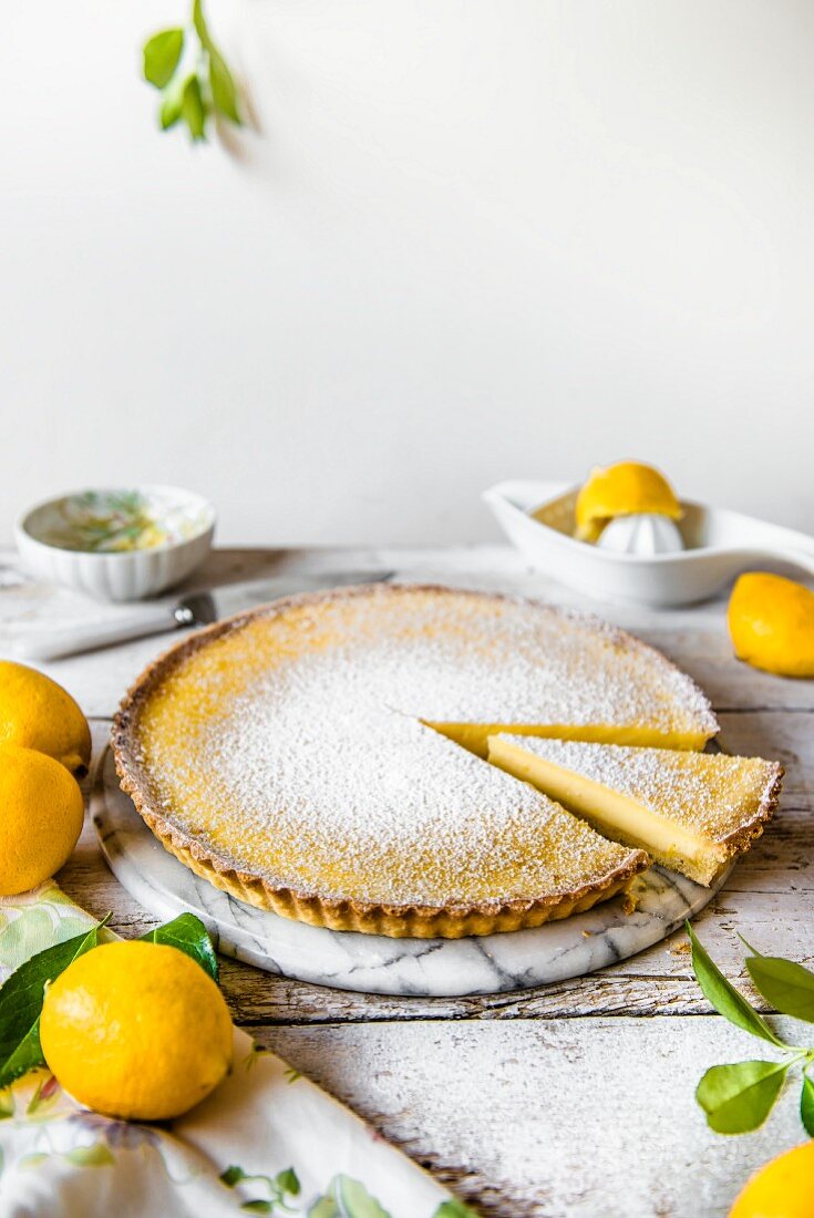 A lemon tart dusted with icing sugar on a marble plate with one slice removed