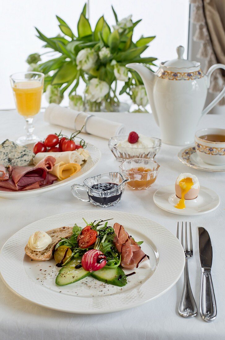 Frühstückstisch mit Prosciutto, Blattsalat, Balsamico, Brot, Butter, Gurke, Radieschen, weichgekochtem Ei und Käseplatte