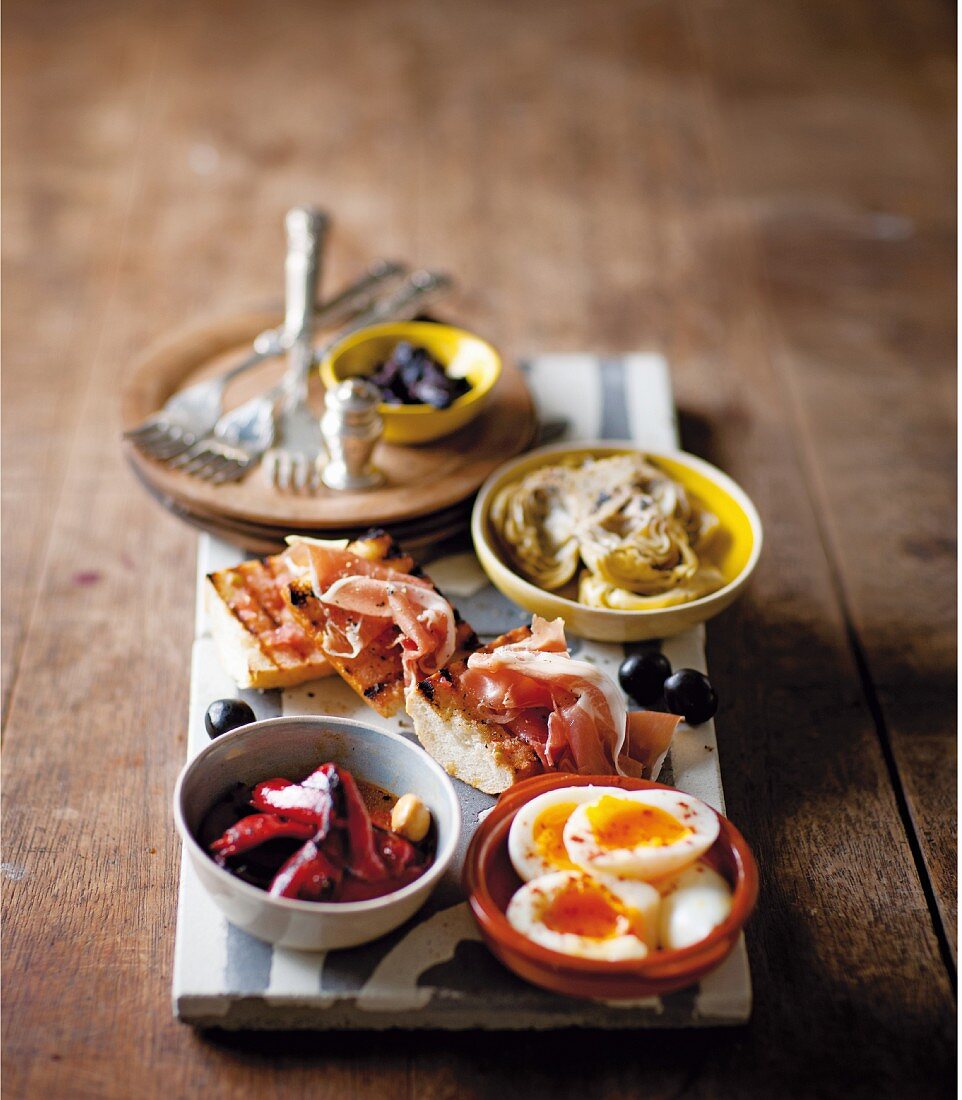Tapas mit marinierten Artischocken, gegrillten Peperonis, Oliven, Eiern und Schinken-Tomaten-Baguette