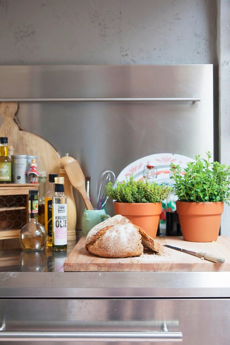 Holzschneidebrett mit Brot und Küchenkräuter in Tontöpfen auf Edelstahl Arbeitsfläche