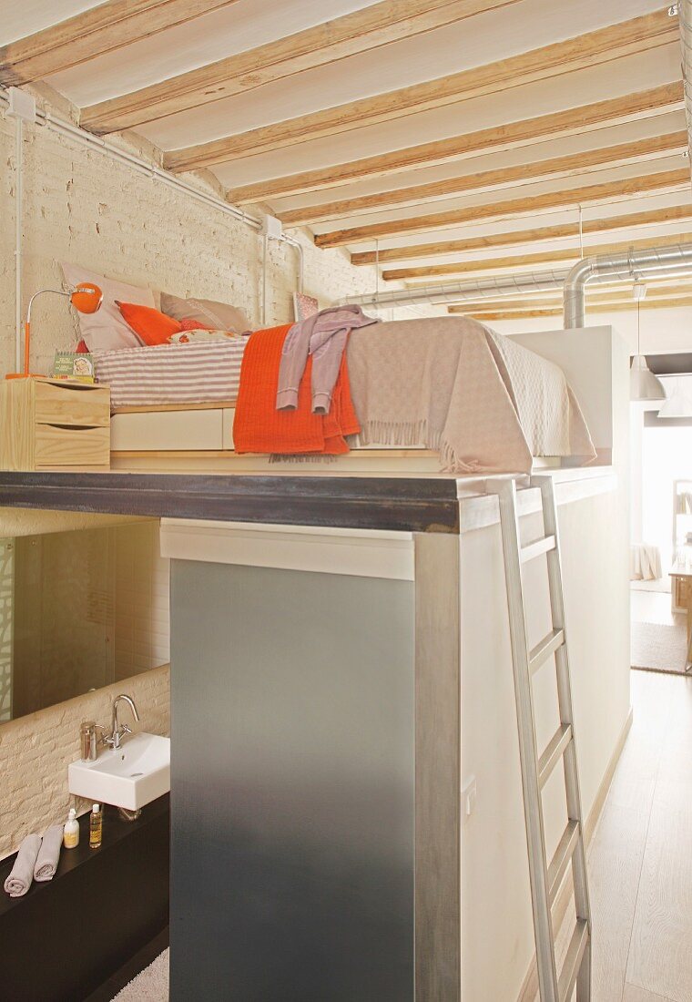 Schlafbereich mit Bett und Nachtkästchen auf eingebautem Kubus mit Metallleiter, darunter Bad in Loftwohnung