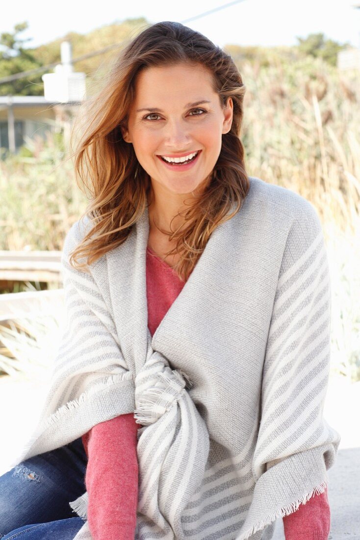 A blonde woman wearing a pink jumper and jeans with a striped blanket over her shoulders