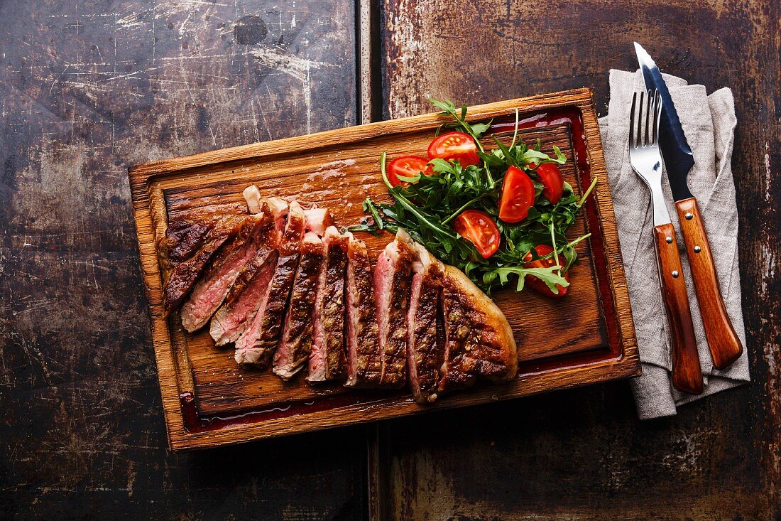 Gegrilltes Striploin Steak in Scheiben geschnitten und Rucola-Tomaten-Salat auf Holzbrett