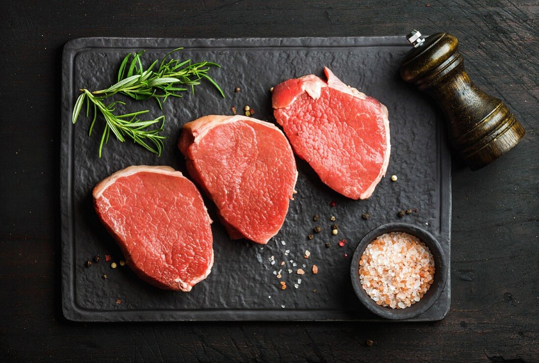 Rohe Rindersteaks (beef eye round steaks) mit Gewürzen und Rosmarin auf schwarzer Schieferplatte