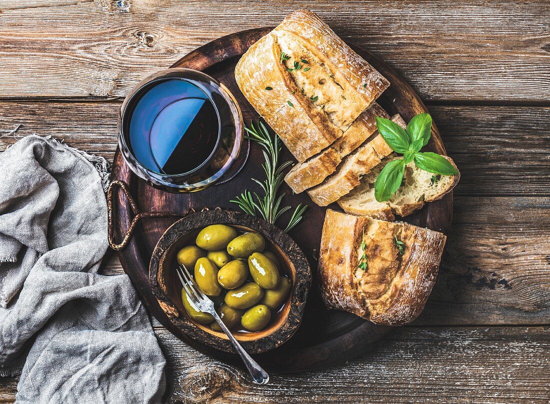 Ein Glas Rotwein, frisch gebackene Ciabatta und grüne Oliven auf Holztablett