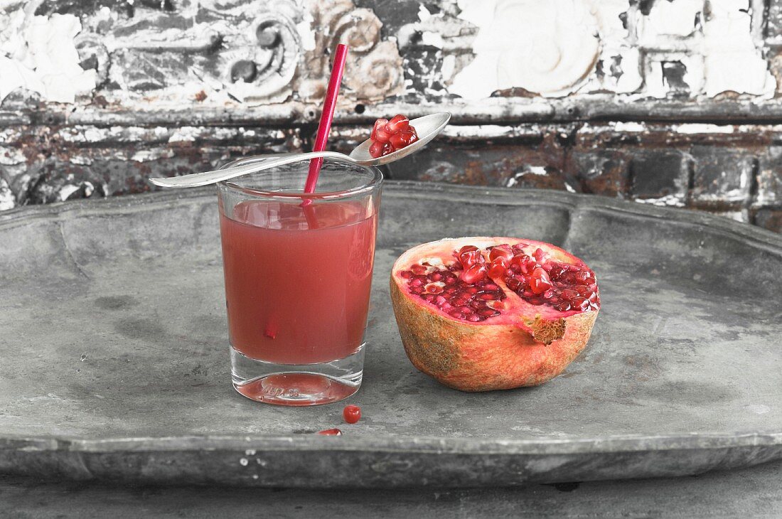 Pomegranate juice in a glass with a straw and spoon next to a pomegranate sliced in half
