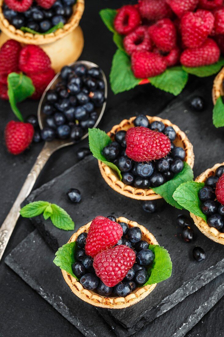 Törtchen mit Heidelbeeren, Himbeeren und Minzblättern auf Schieferuntergrund