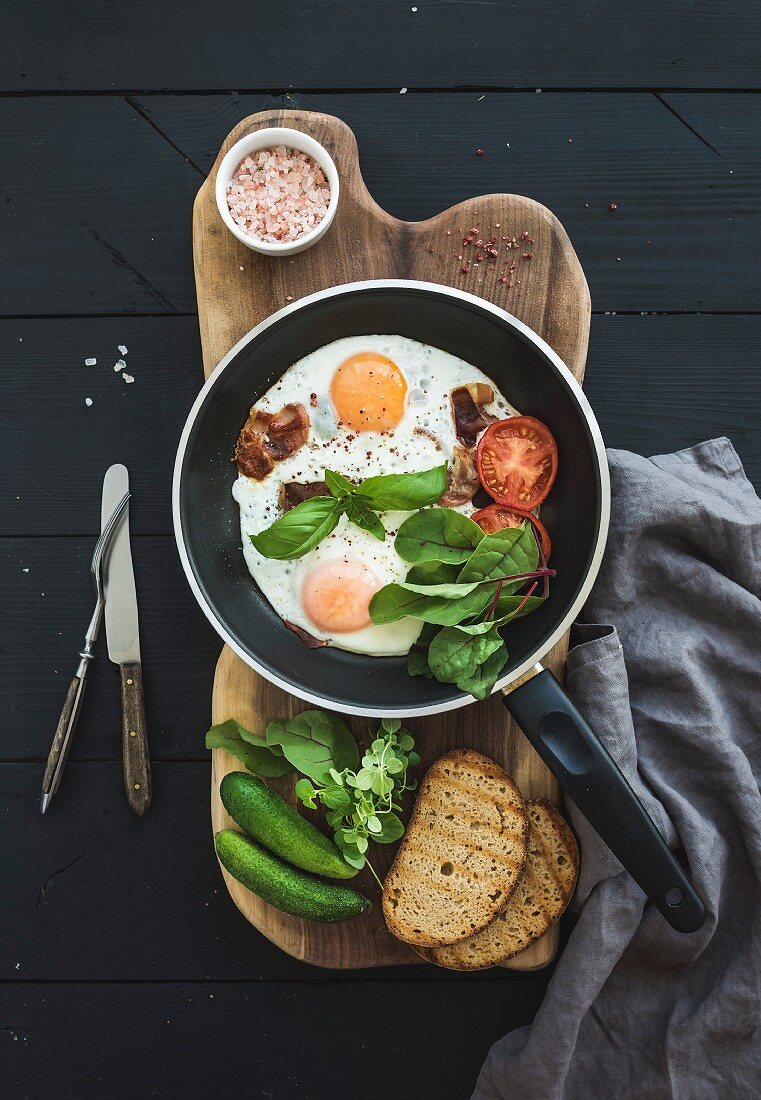 Pan of fried eggs, bacon, cherry tomatoes and fresh herbs with bread on wooden board over dark wooden background