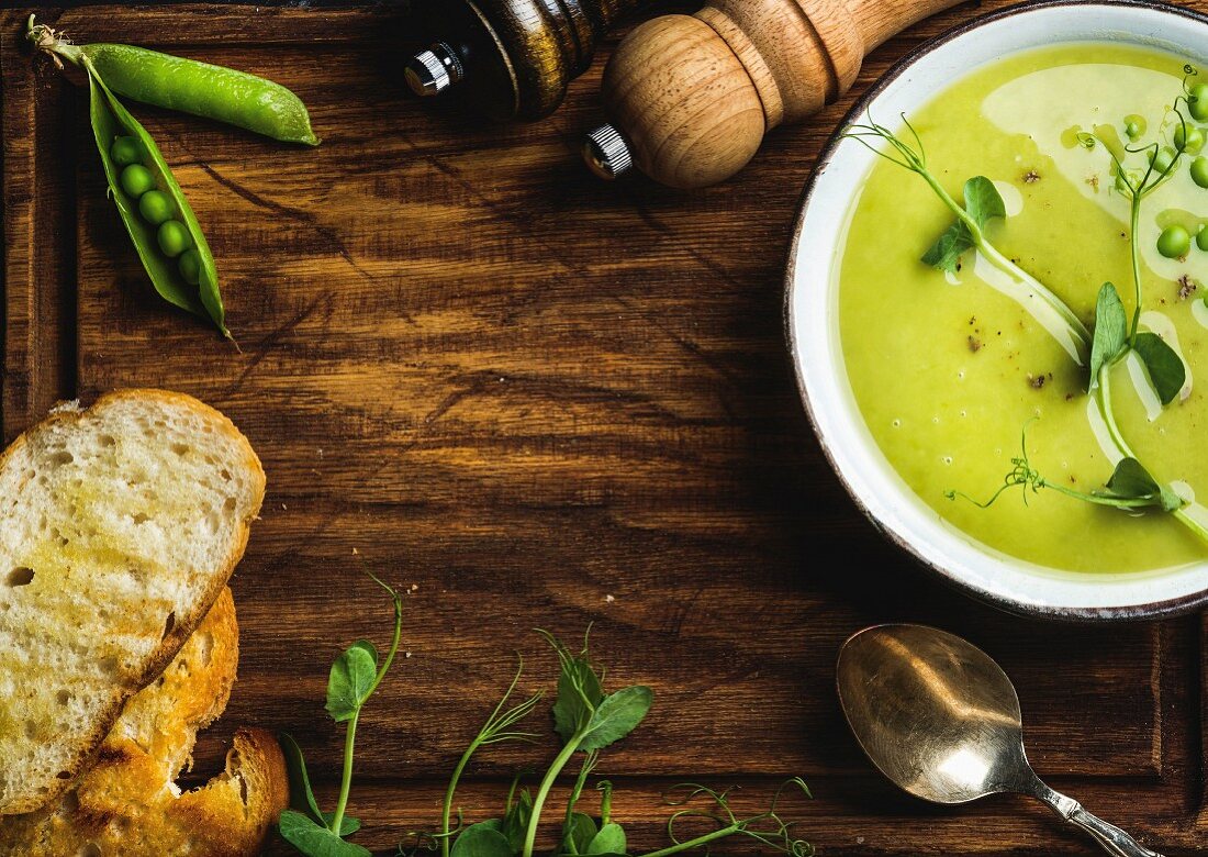 Erbsencremesuppe in weisser Schüssel, dazu Brot auf rustikalem Holzbrett