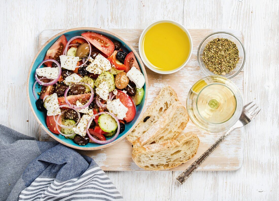 Griechischer Salat mit Olivenöl, Brot, Oregano und Glas Weißwein auf Holzbrett