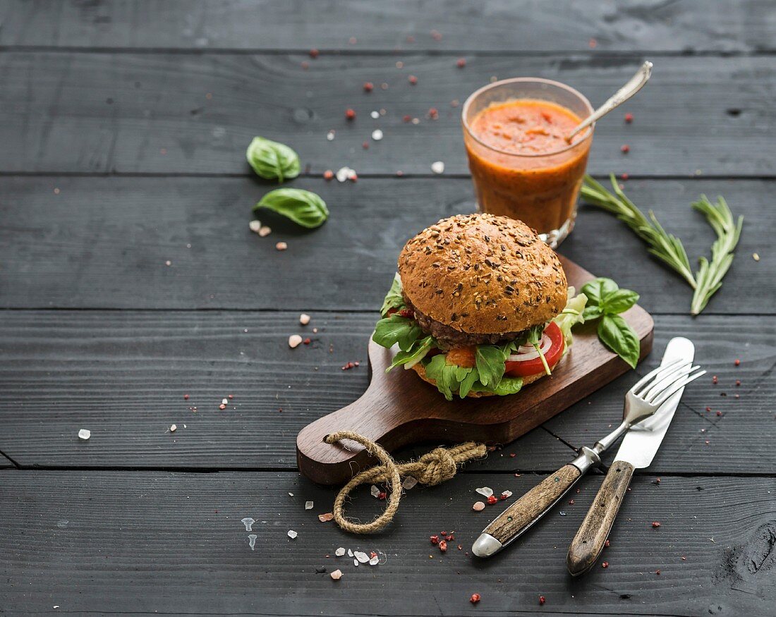 Fresh homemade burger with vegetables and tomato sauce on wooden serving board over dark wooden background