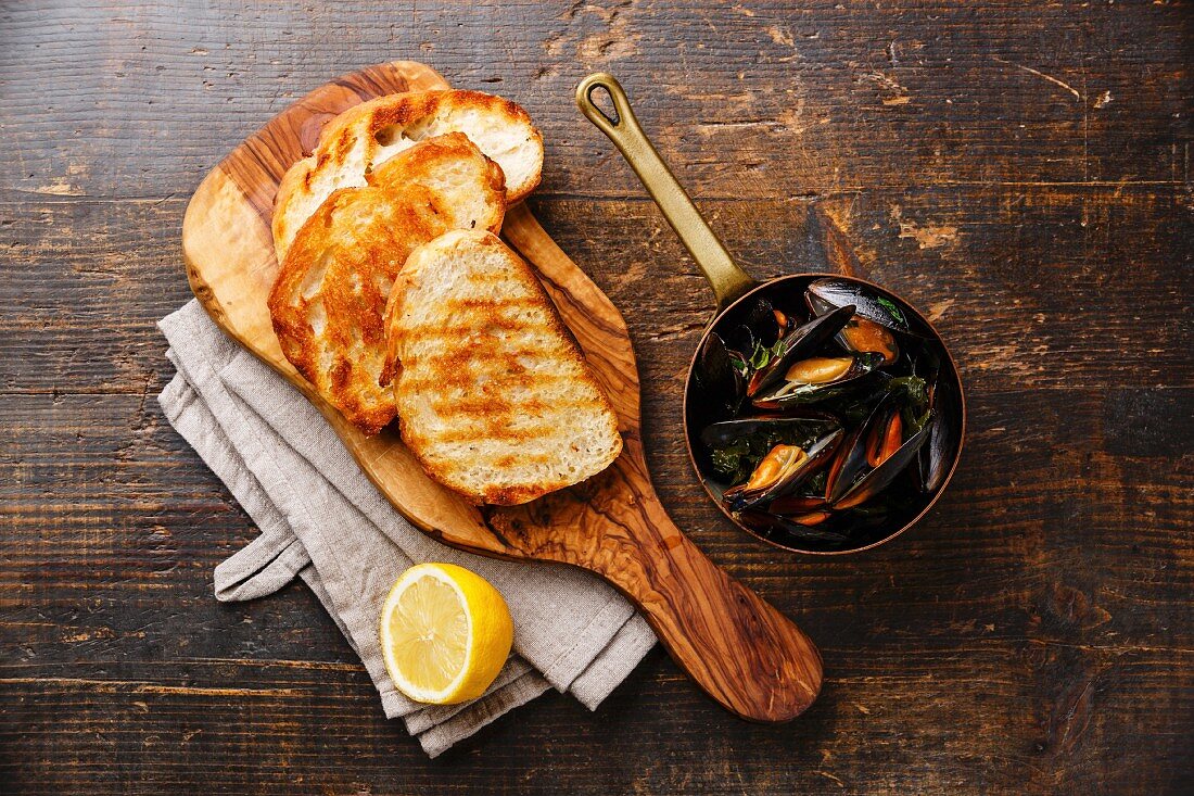 Mussels in copper pot, bread toasts and lemon on wooden background
