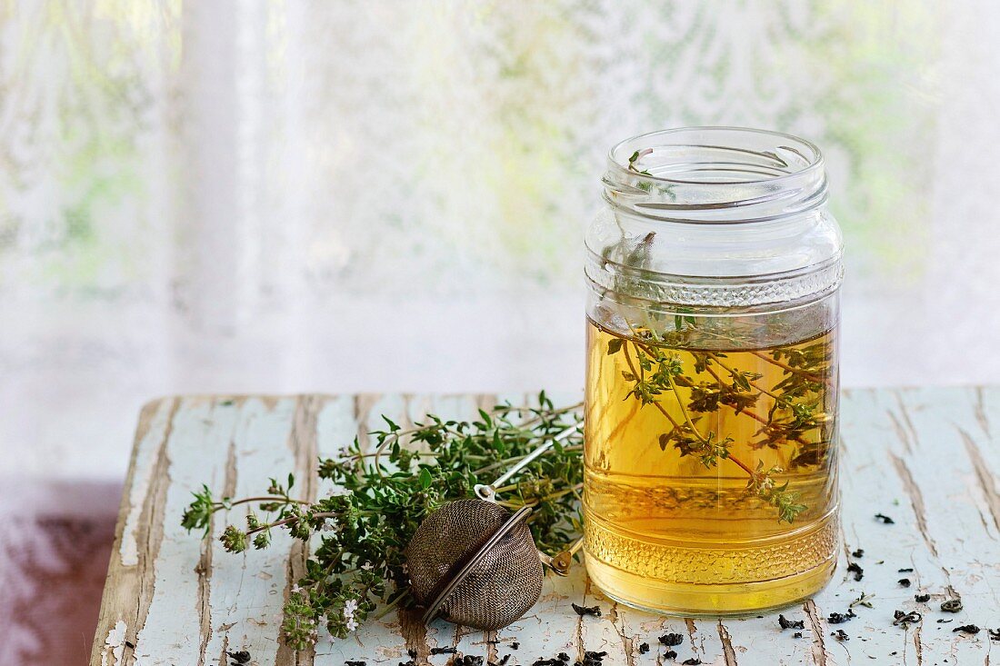 Glass jar of hot herbal tea with bunch of fresh thyme, served with vintage tea-strainer on old wooden stool