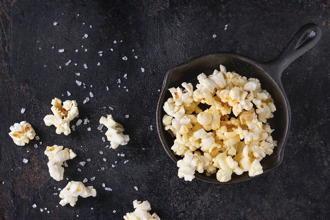 Prepared salted popcorn served with sea salt in small iron-cast pan over black iron textured background