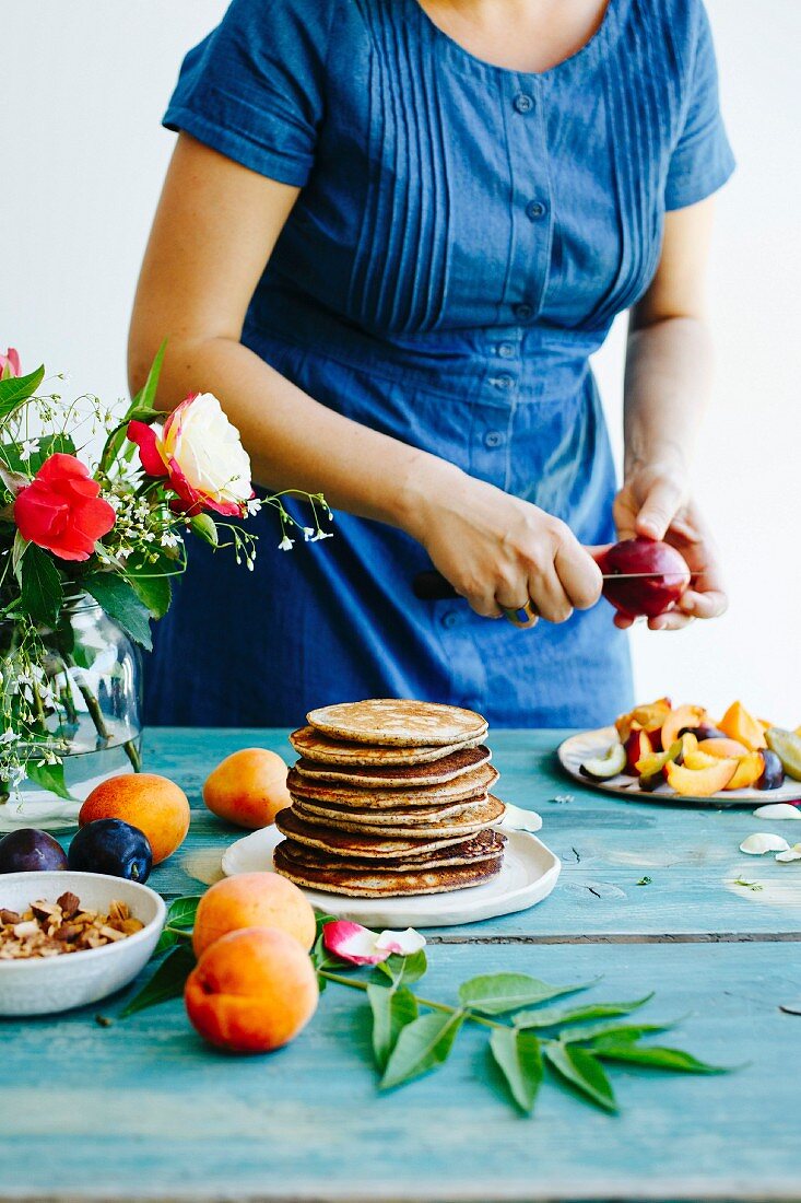 Gestapelte Pancakes umgeben von Früchten auf türkis Holztisch, Mädchen in Jeanskleid im Hintergrund