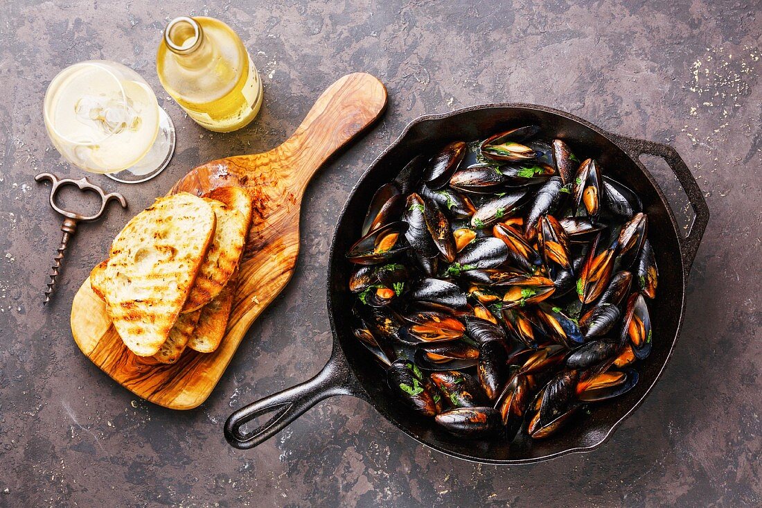 Miesmuscheln in einer Pfanne, getoastetes Brot und Weißwein auf dunklem Untergrund