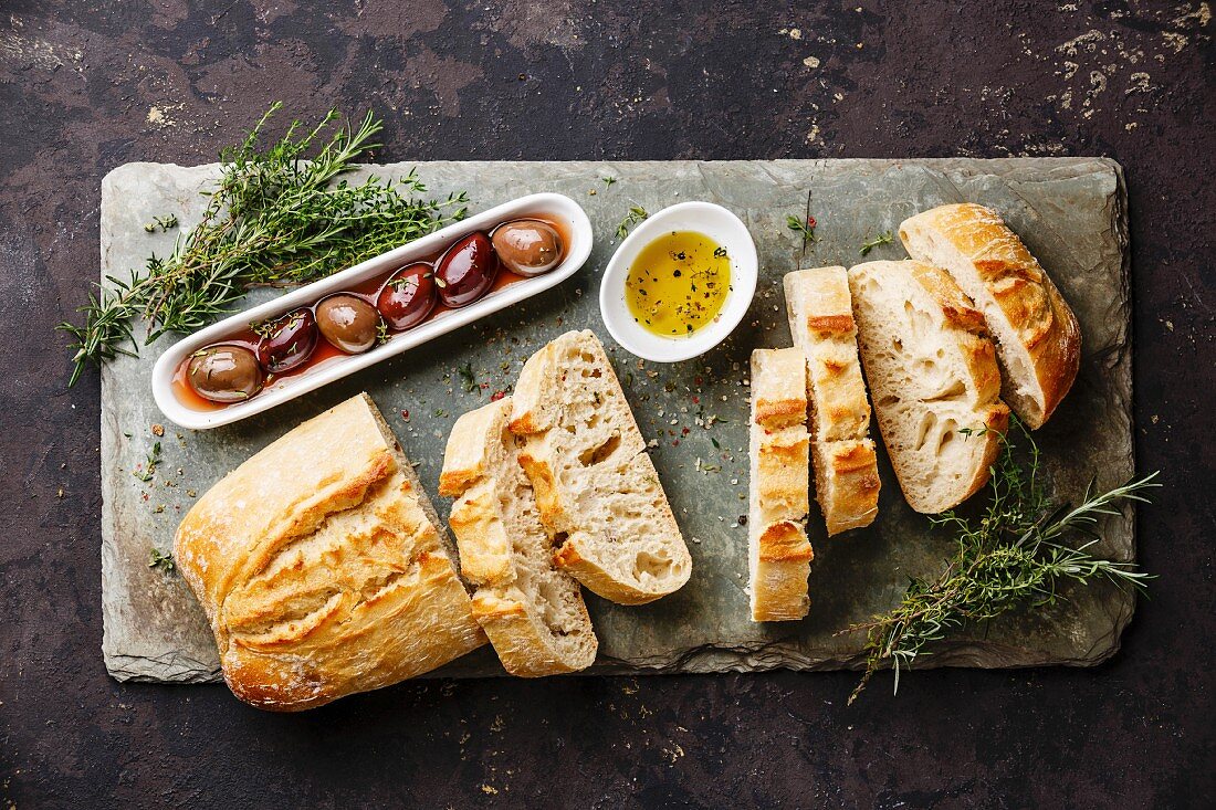 Fresh Ciabatta bread cut in slices on stone slate board with olives, oil and herbs on dark background