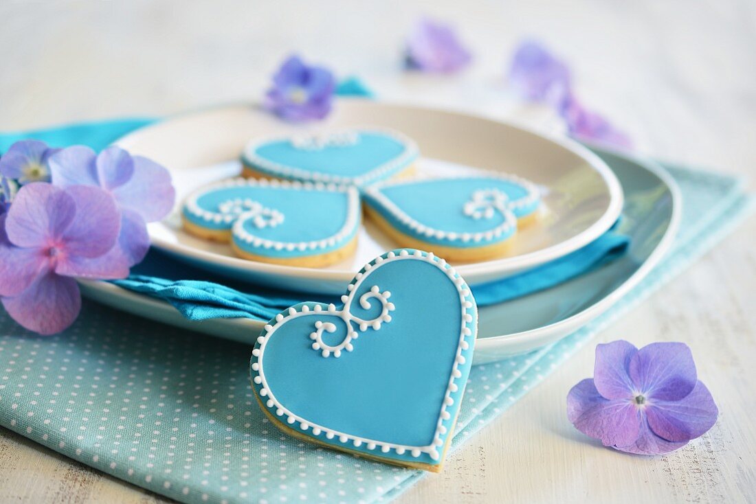 Kekse in Herzform mit blauem und weißem Zuckerguss dekoriert, auf Teller und mit Blumen serviert