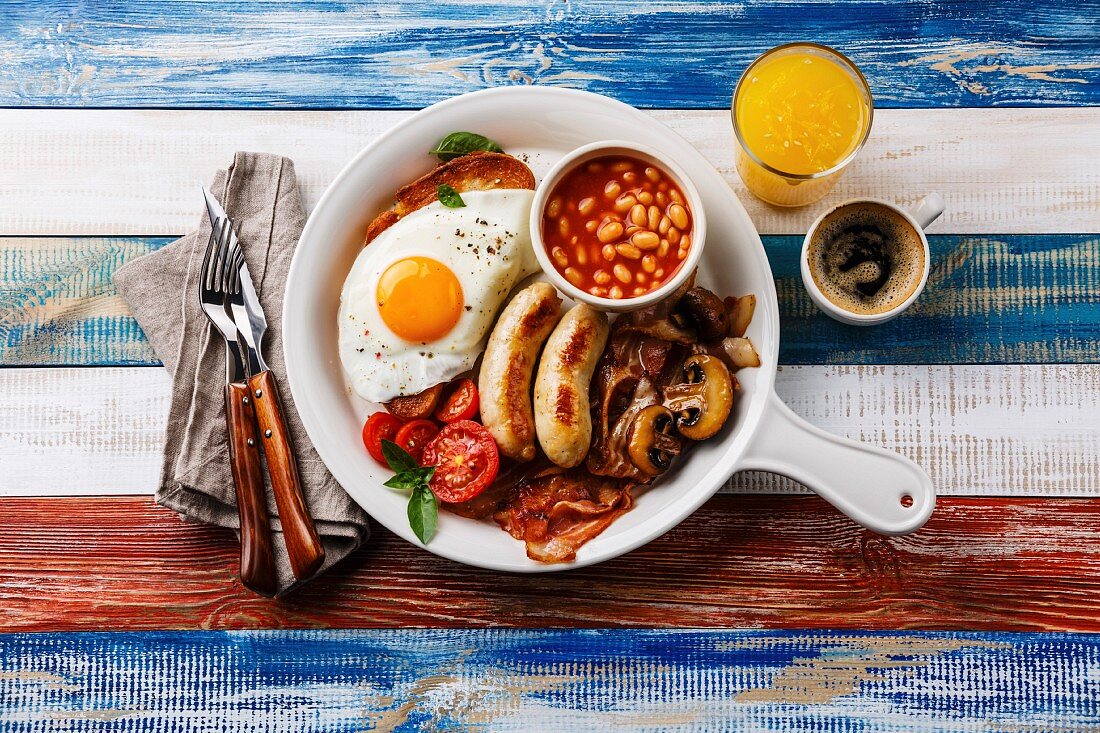 English breakfast in white pan with fried eggs, sausages, bacon, beans, toast, orange fresh and coffee on wooden background