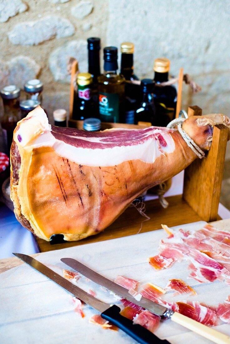 A whole air dried ham on a wooden stand with knives, bottles and a chopping board