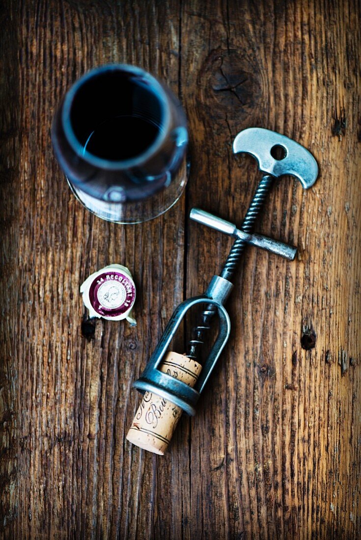 An antique corkscrew with a cork, and a glass of red wine on a wooden surface