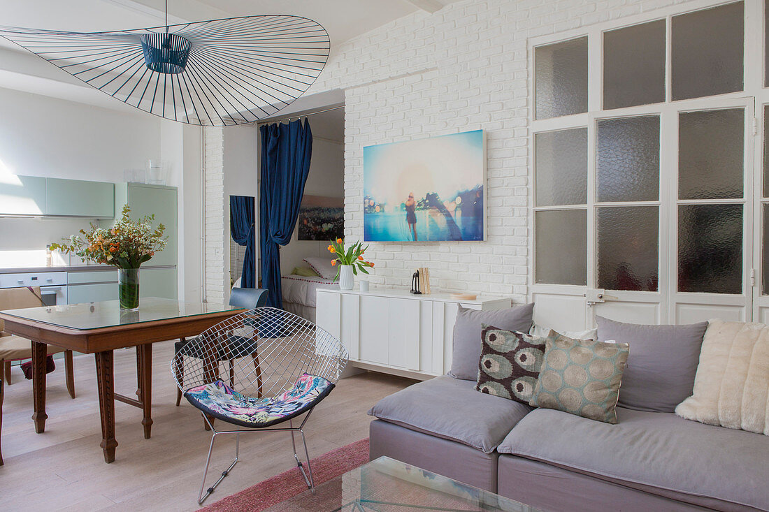 Scatter cushions on grey sofa, interior door with glass inserts, classic chair, dining table and kitchen in open-plan apartment