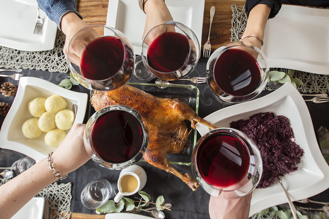 People raising their glasses of red wine over a roast goose with red cabbage and potato dumpling