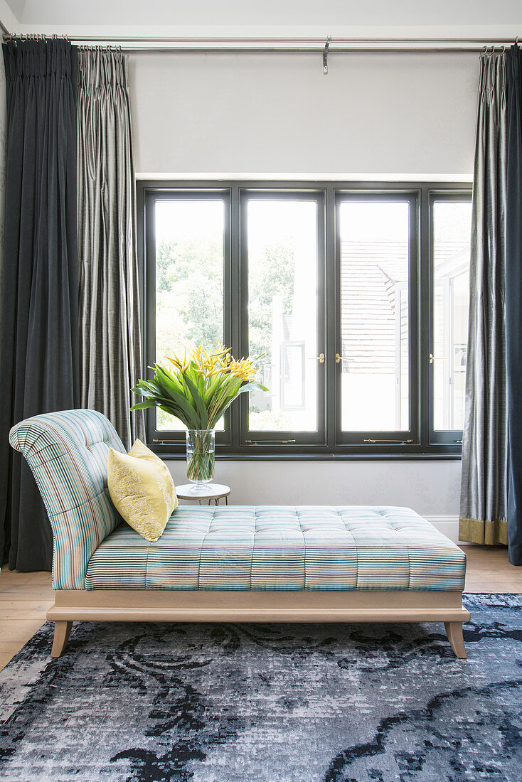 Stylish chaise longue and flowers on side table in front of window in bedroom