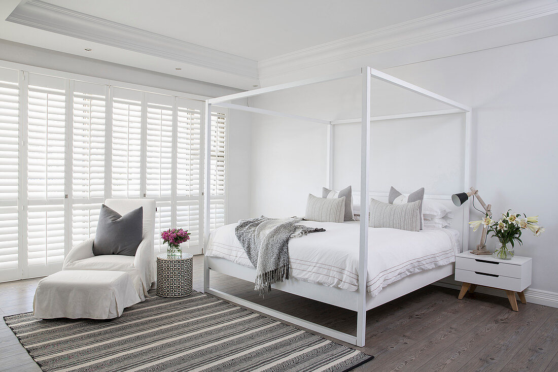 Simple four-poster bed and armchair with footstool in white bedroom