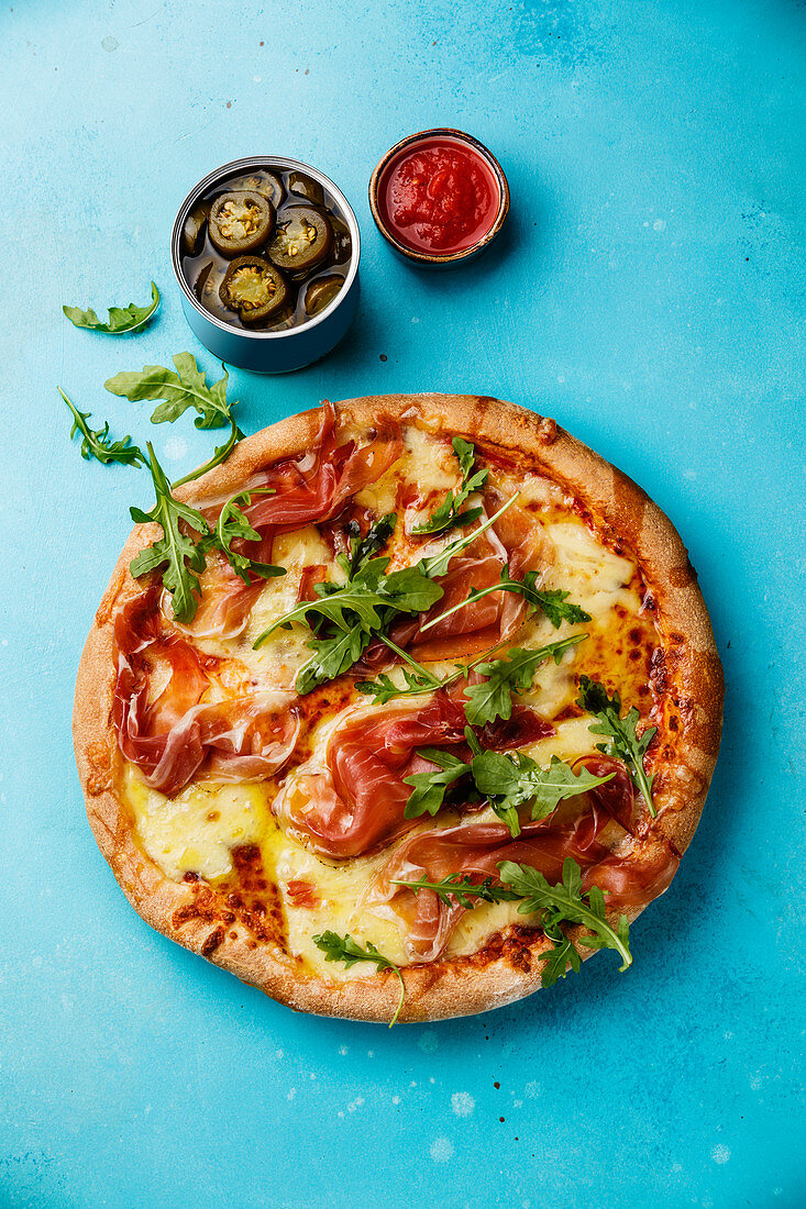 Pizza with mozzarella cheese, prosciutto and arugula leaves on blue background