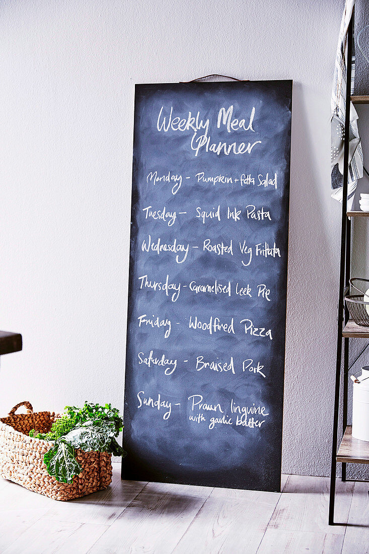 DIY-Kreidetafel mit Wochenmenü an die Wand gelehnt, davor Korb mit Gemüse