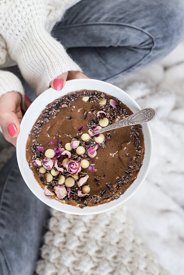 A woman holding a smoothie bowl with chocolate, dried roses and cocoa nibs