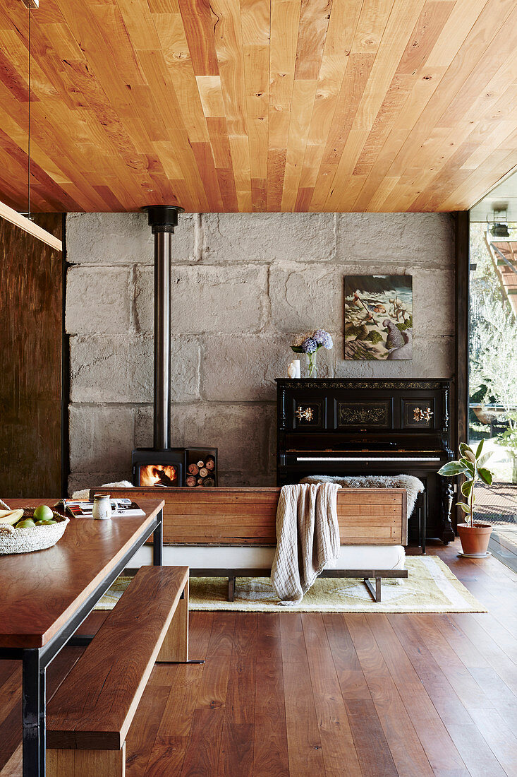 Dining area in an open living room with concrete wall and wooden ceiling, in the background Swedish stove and piano