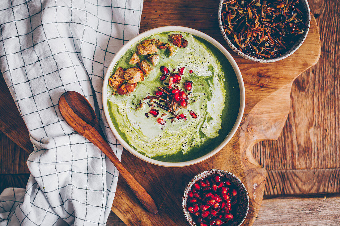 Spinach soup with wild rice, cream, croutons and pomegranate seeds on a wooden board with a tea towel and a spoon