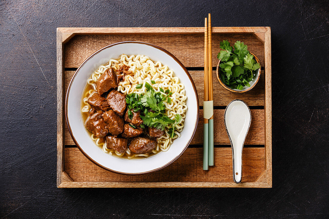 Slow cooked Beef meat with Asian noodles in broth in wooden tray on dark background