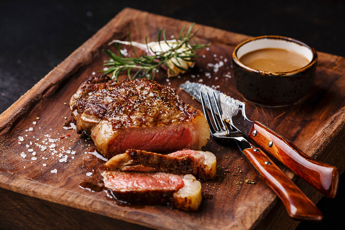 Gegrilltes New York Striploin Steak mit Pfeffersauce und Besteck auf Holzbrett