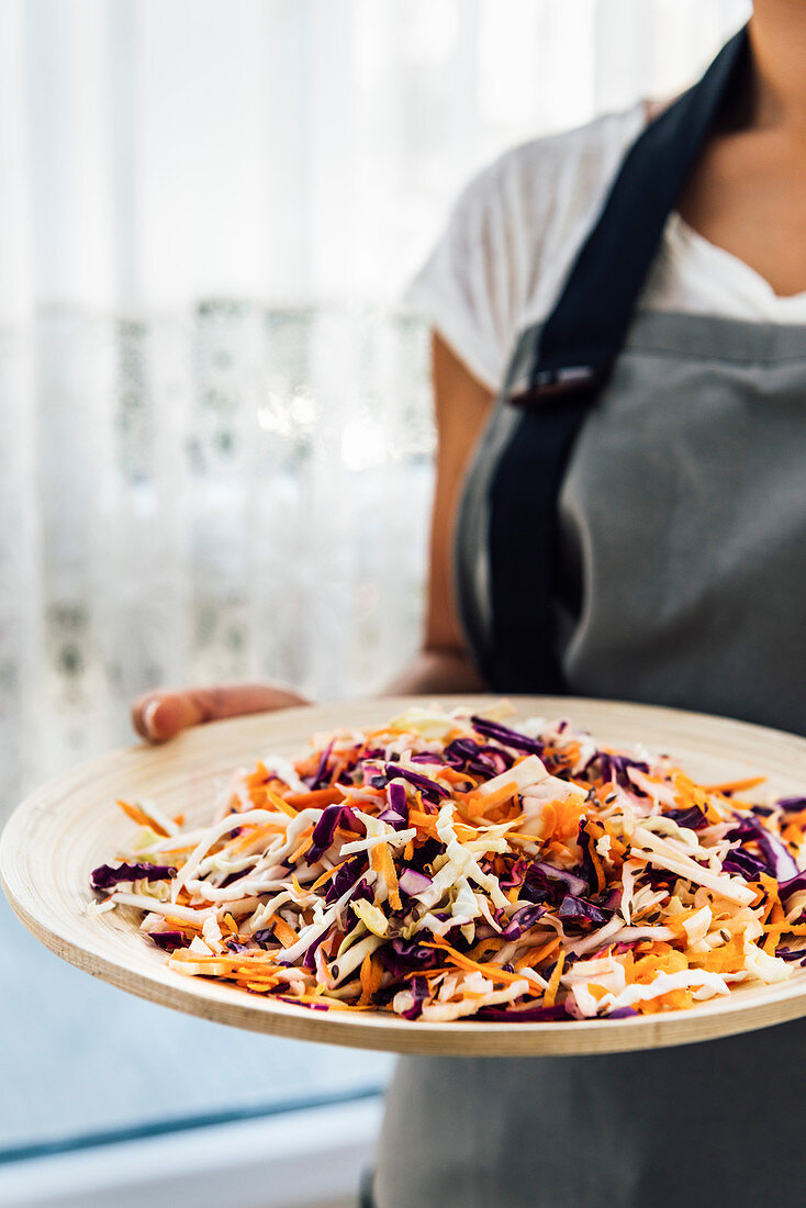 A woman holding a wooden bowl with coleslaw made with shredded carrot, white and purple cabbage