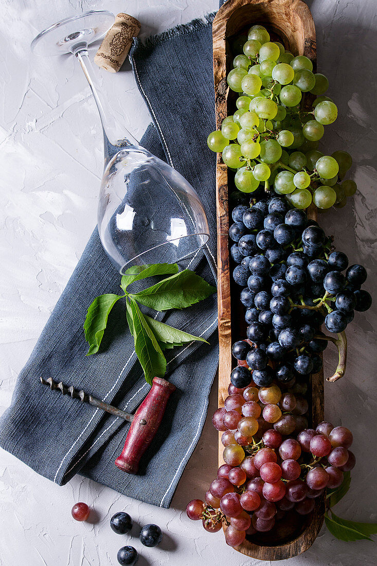 Variety of three type fresh ripe grapes dark blue, red and green in wooden bowl with empty laying wine glass, old corkscrew and green leaves