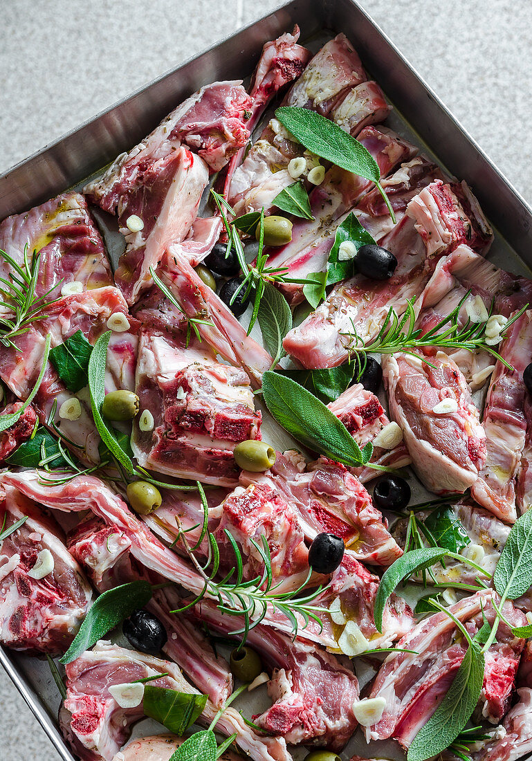 Rohe Stücke vom Lamm mit Knoblauch, Oliven, Salbei, Rosmarin und Lorbeer auf Backblech