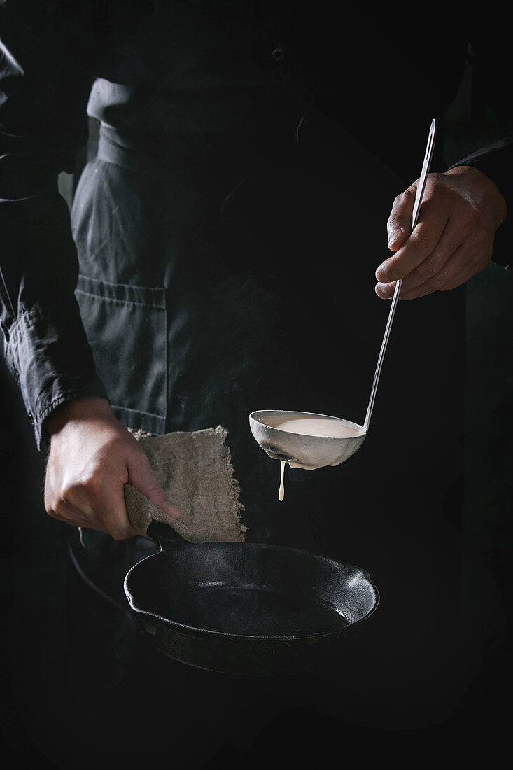 Man chef in black apron pouring dough from ladle for cooking pancakes in cast-iron pan