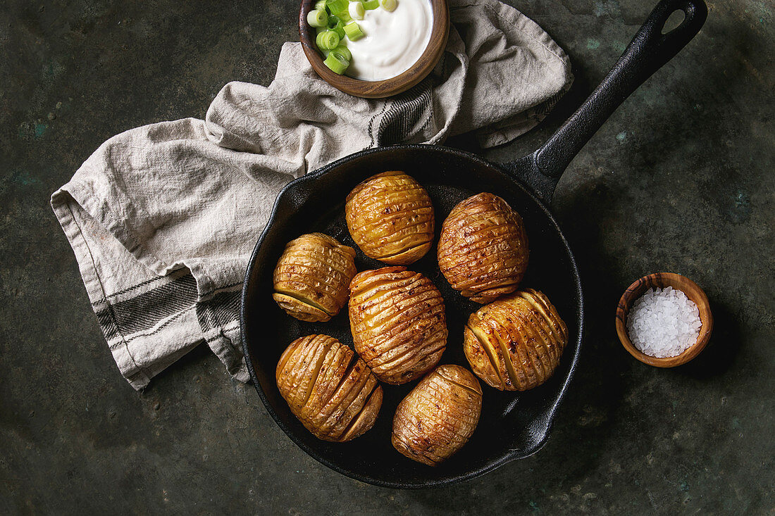 Accordion hasselback baked potatoes served in cast-iron pan with sea salt, cream-fresh and green spring onion on textile napkin