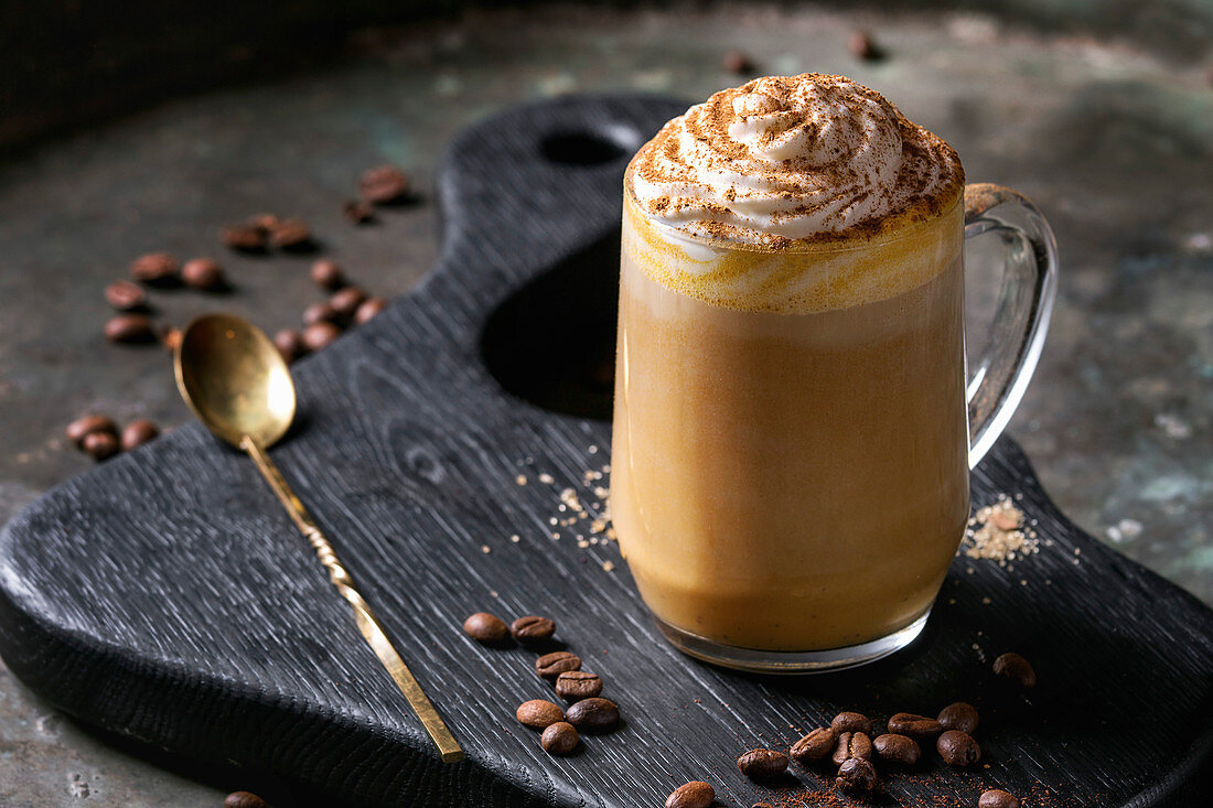 Glass of spicy pumpkin latte with whipped cream and cinnamon standing on black serving board