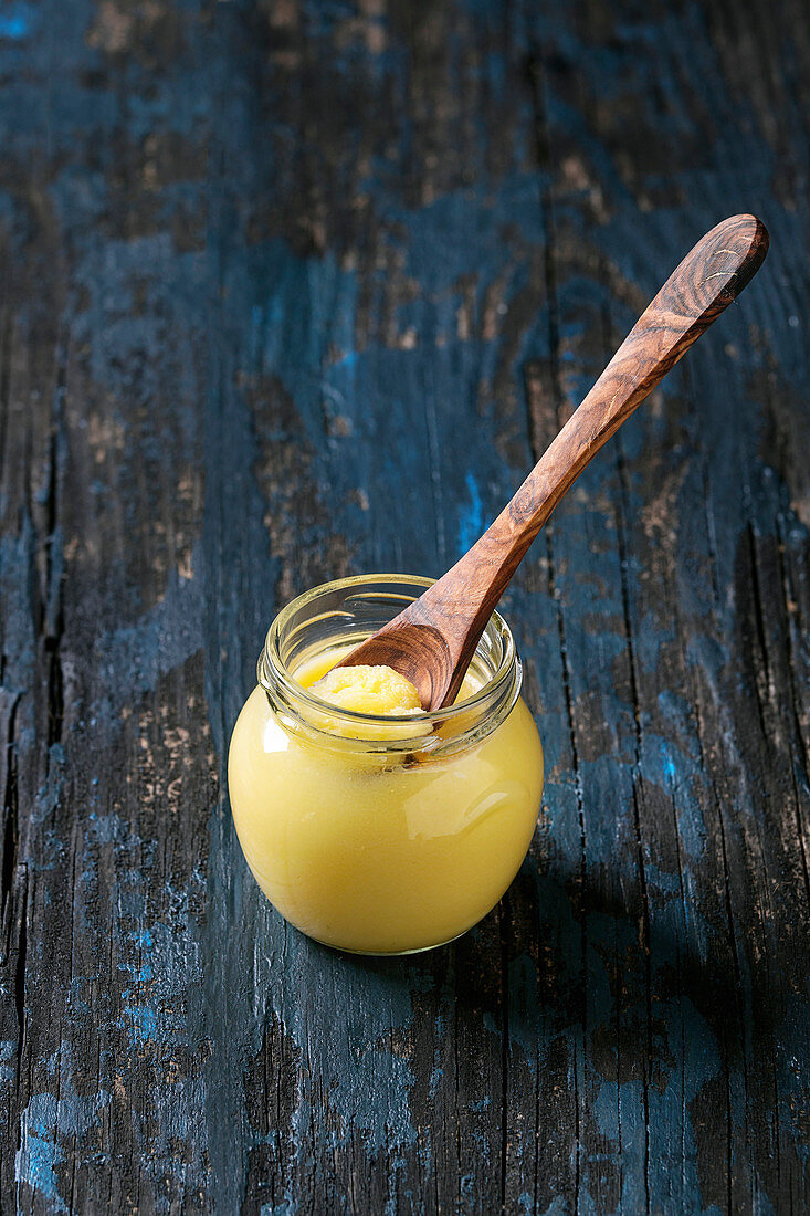 Glass of drawn ghee butter standing with wood spoon over old dark blue wooden background