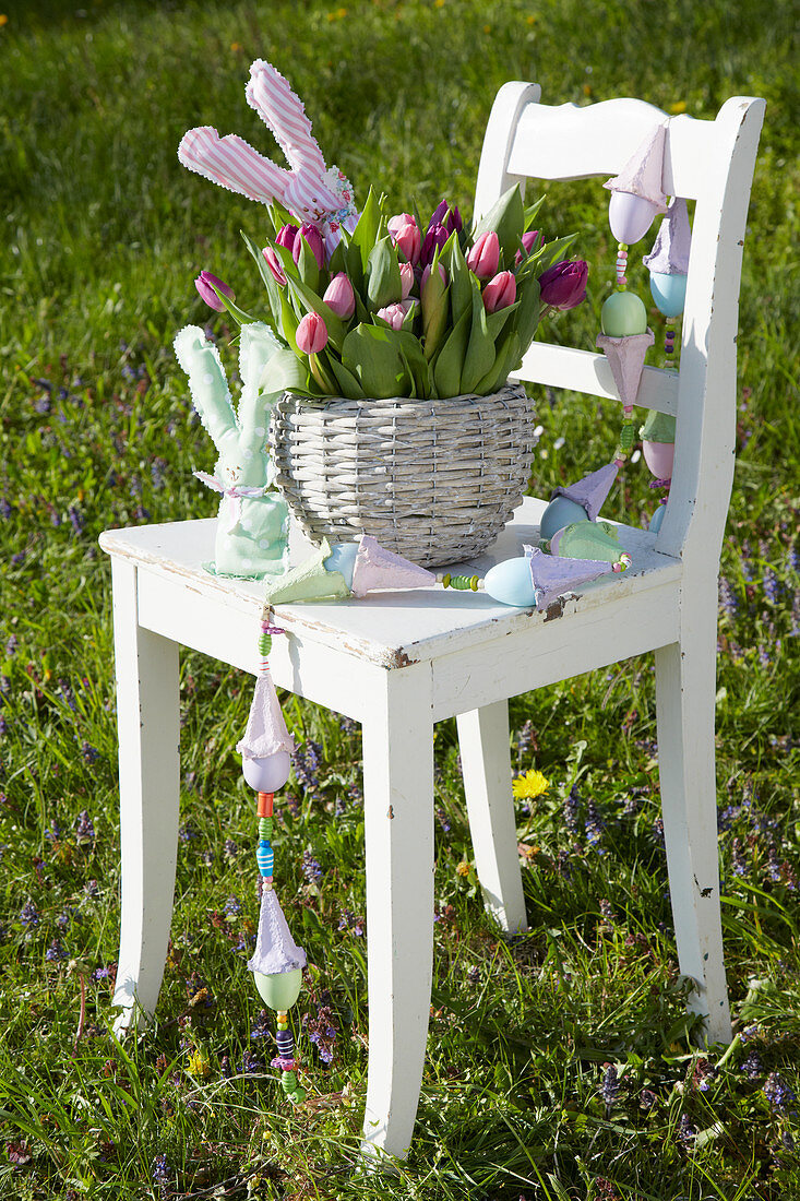 Bunch of tulips and hand-made fabric bunnies in basket and garland of eggs on chair in garden